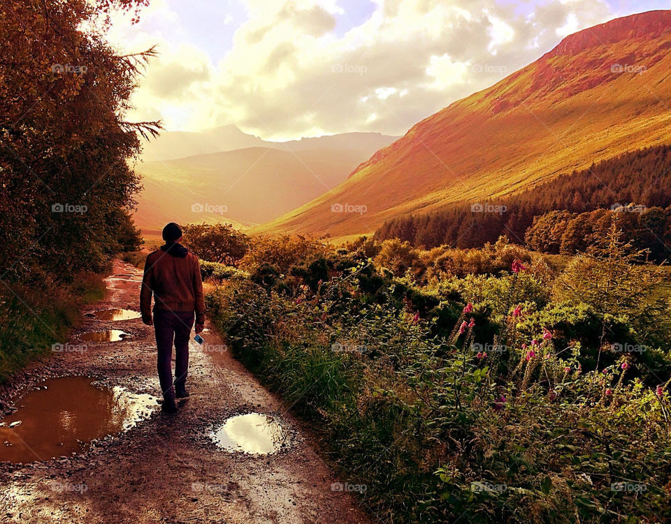 Morning walk on the Isle of Arran, hazy sunshine breaks through the cloud to light up the hills as a man dodges puddles on the trail