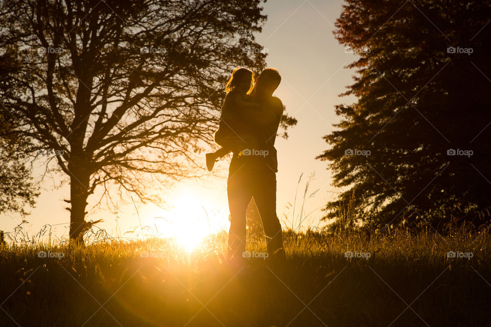Dad and his girl standing in a field at sunset enjoying quality time and having fun. Trees and golden sun with people silhouetted.