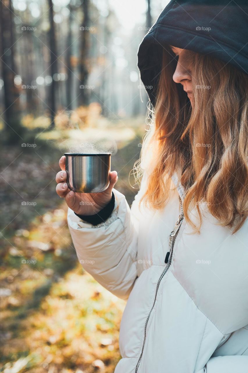 Woman in a hood having break during autumn trip holding cup with hot drink from thermos flask on autumn cold day. Active girl wandering in a forest actively spending time