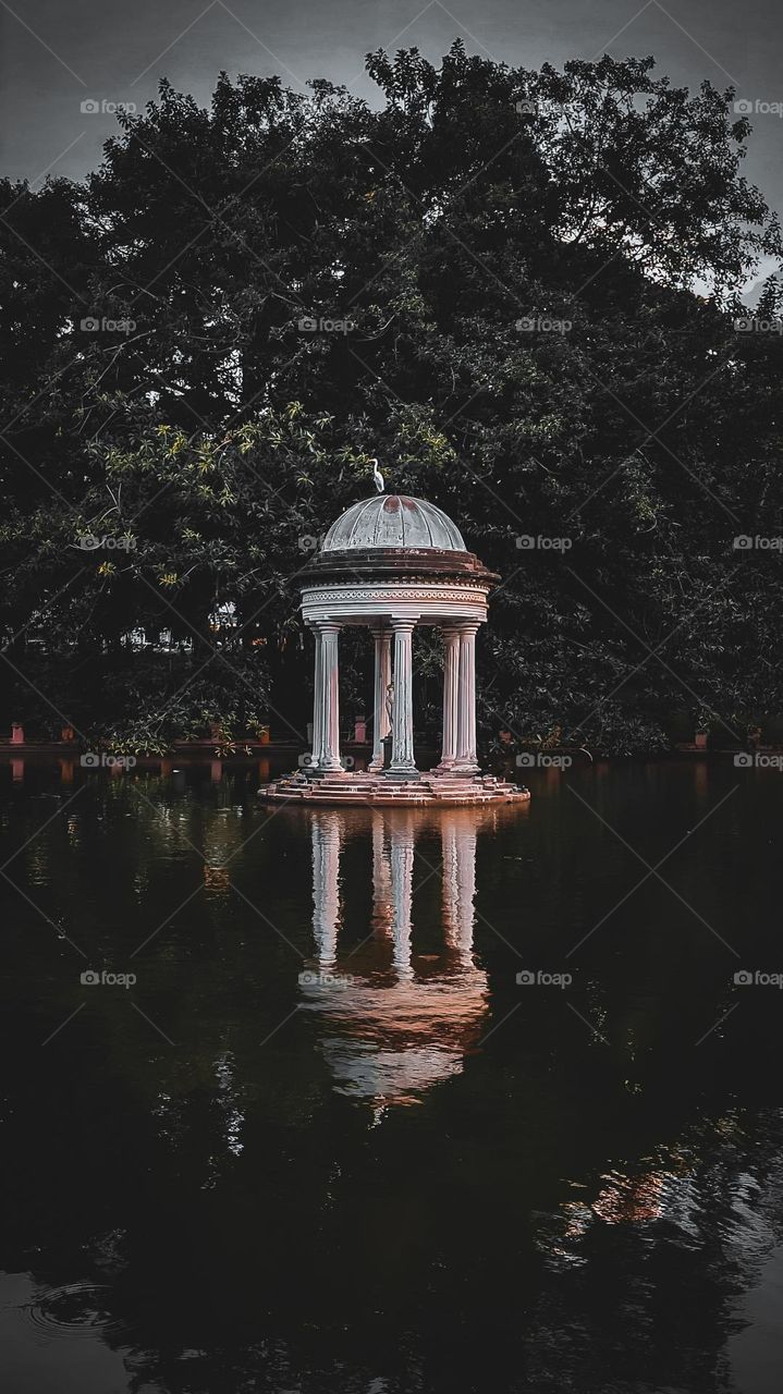 A small classical pavilion reflects on calm waters, surrounded by dense trees. Soft light adds a mysterious and serene mood, highlighting the elegant architecture against the dark, natural backdrop.