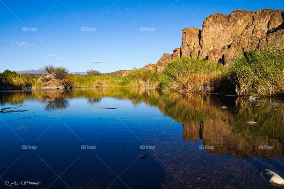 A still pond reflects the beautiful blue sky and golden rock wall in the Arizona desert on a flawless spring day