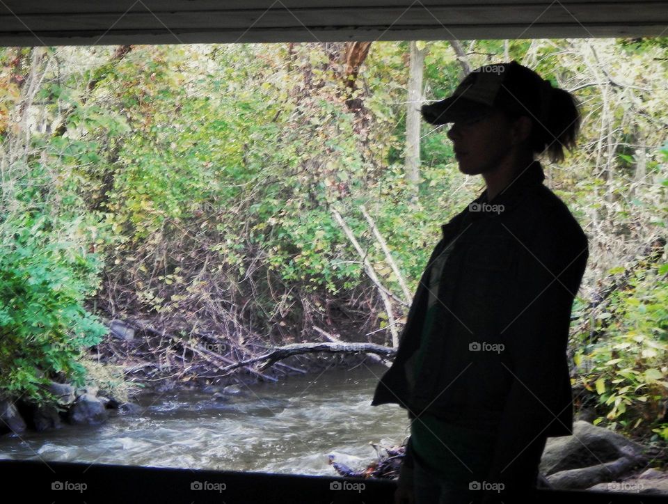 Woman in baseball cap and ponytail stands near a river in Michigan under a bridge. The woman is in a shadow and behind her is the river and green trees plants and rocks