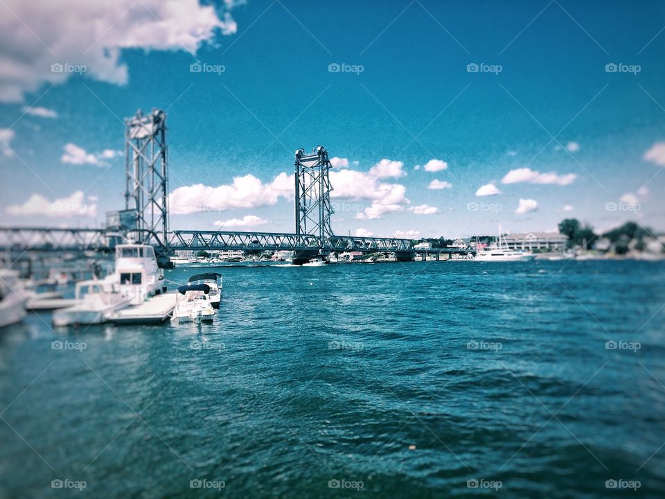 The blue ocean view of Portsmouth, New Hampshire with a bright cloudy sky, boats, and a bridge. 
