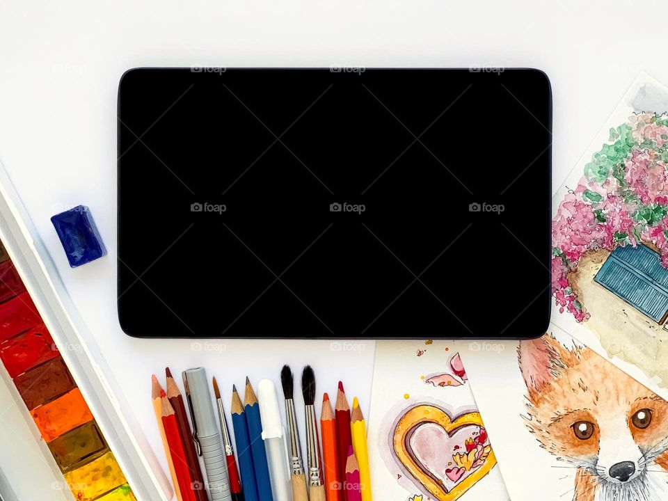 Tablet with black screen, watercolor sketches, paints, brushes, colored pencils, liner on white table. 