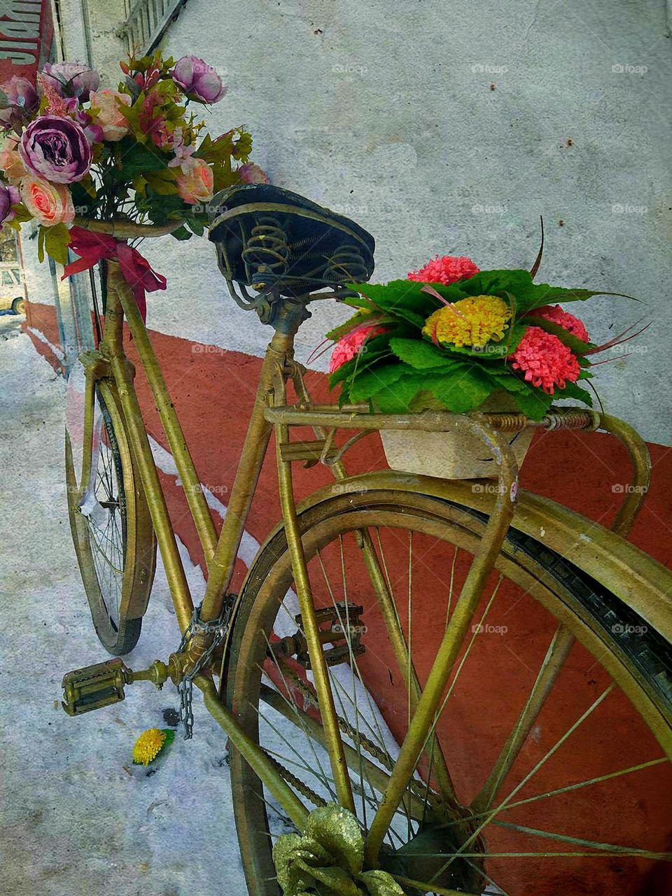 Bicycles. Near the wall is a bicycle painted in golden color. Flowers are attached to the handlebars of a bicycle. There is a flower basket on the rear frame of the bicycle. Bicycle stands on white snow