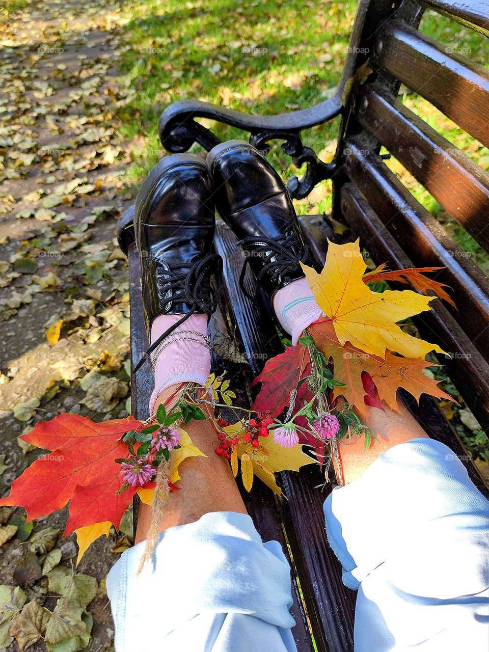 Autumn. A wooden bench stands among autumn trees. On the bench are legs in blue trousers, pink socks and black shoes. Bouquets of multi-colored maple leaves, pink clover and red rowan stick out of the socks.