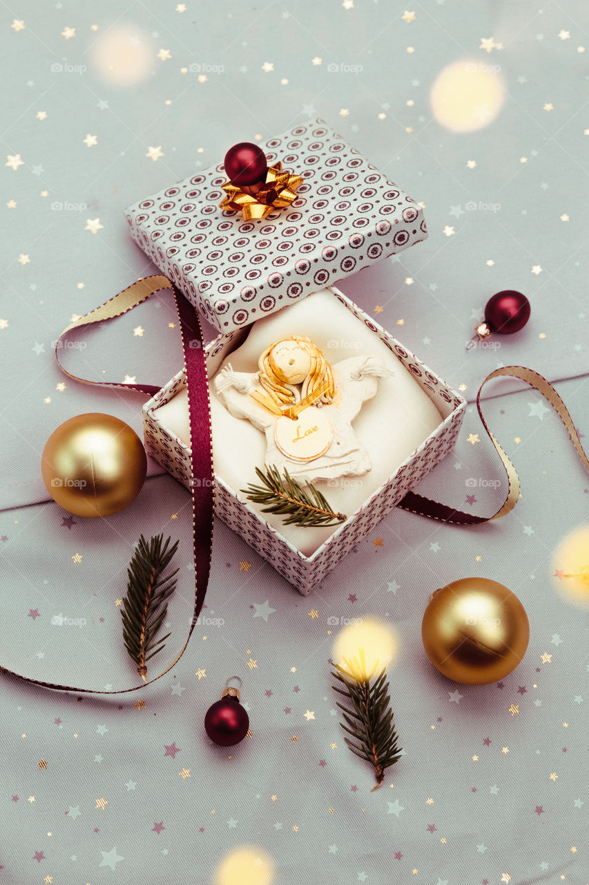 Wrapping Christmas gift into box. Decorating using red ribbon, red Christmas balls and pine twigs. Blurred Christmas lights. Flat top-down composition. Figure of angel made of earthen with love inscription as Christmas decoration