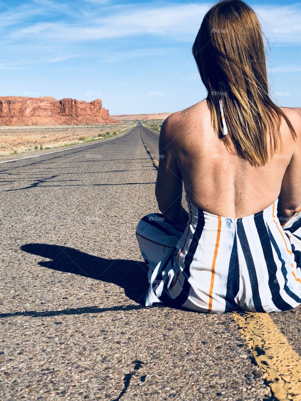 A women sits on the Yello central markings of a highway that cuts throughout the great plains.