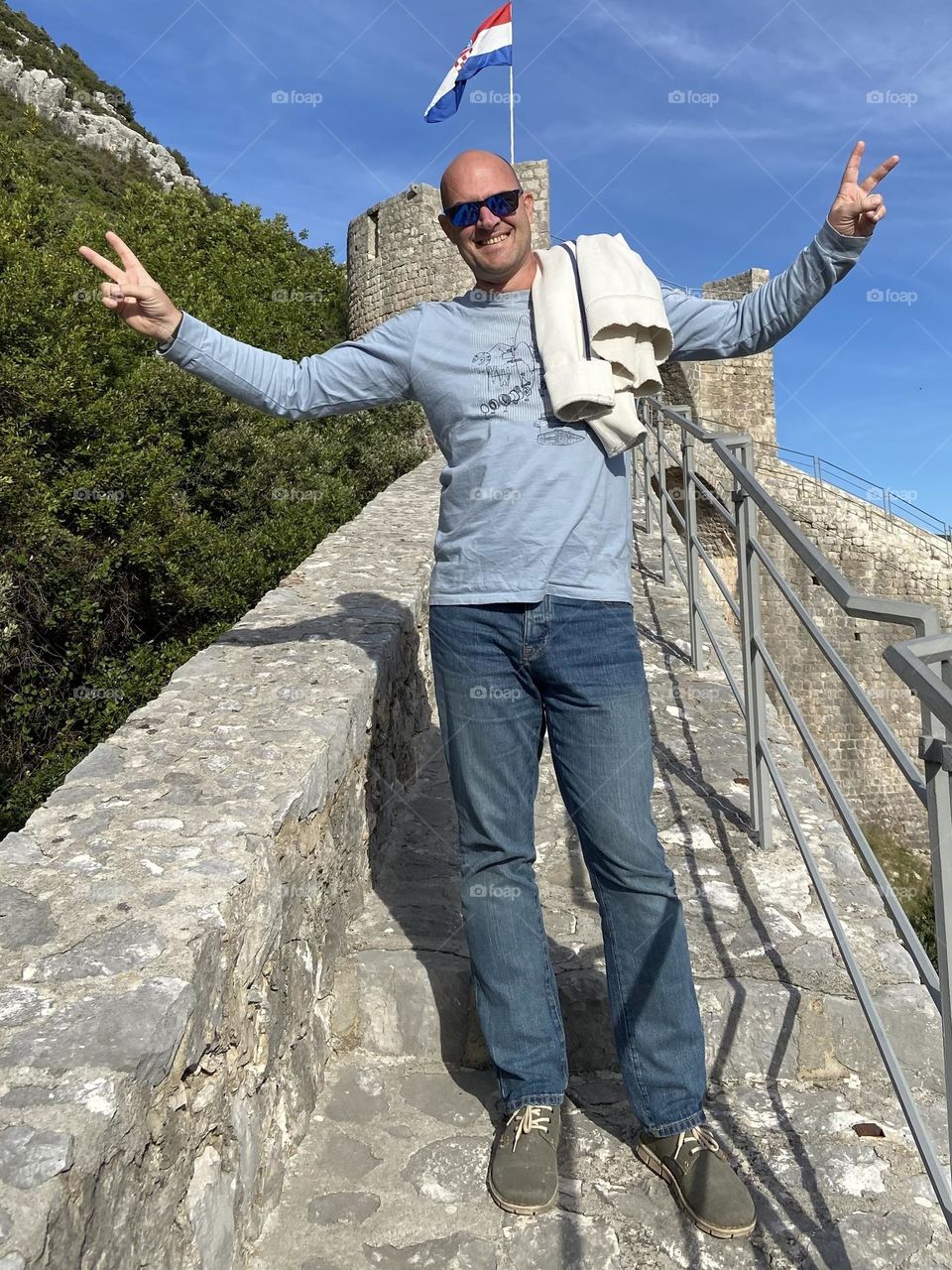 On a hike on the Ston walls in Croatia, a man in a blue shirt and sunglasses is giving peace signs and smiling.