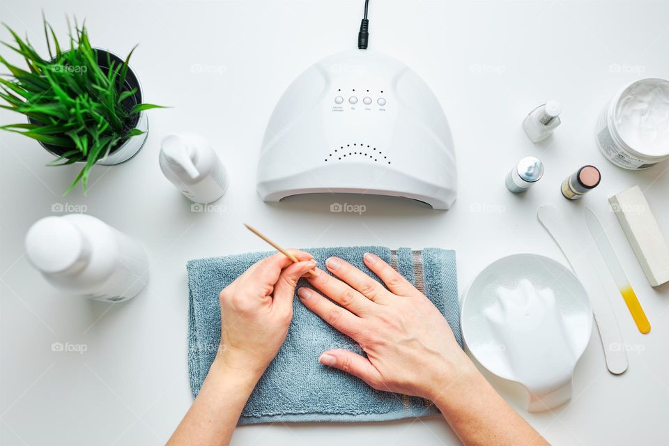 Woman preparing nails to apply gel hybrid polish using UV lamp. Beauty wellness spa treatment concept. Cosmetic products, UV lamp, green leaves on white table. Spa, manicure, skin care concept. Flat lay, overhead view