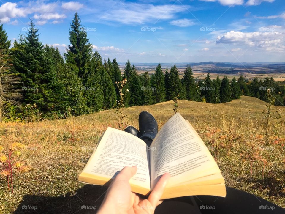 Woman with black boots laying on the grass reading a book surrounded by a beautiful landscape in autumn