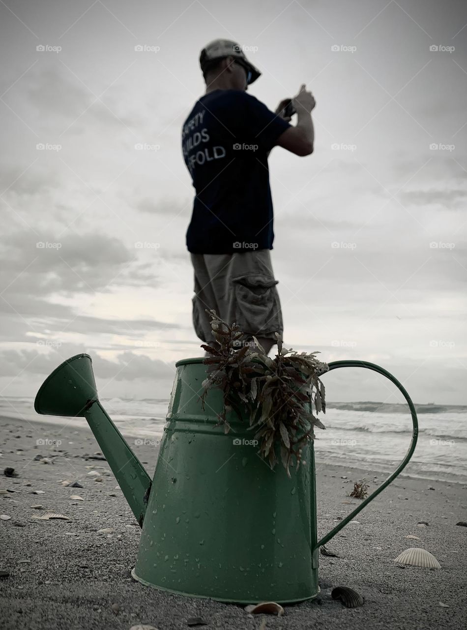 Dad Taking A Picture On The Seashore Who Seems To Come Out Of A Green Watering Can On The Sand With Seashells At The Atlantic Ocean On A Cloudy Day.