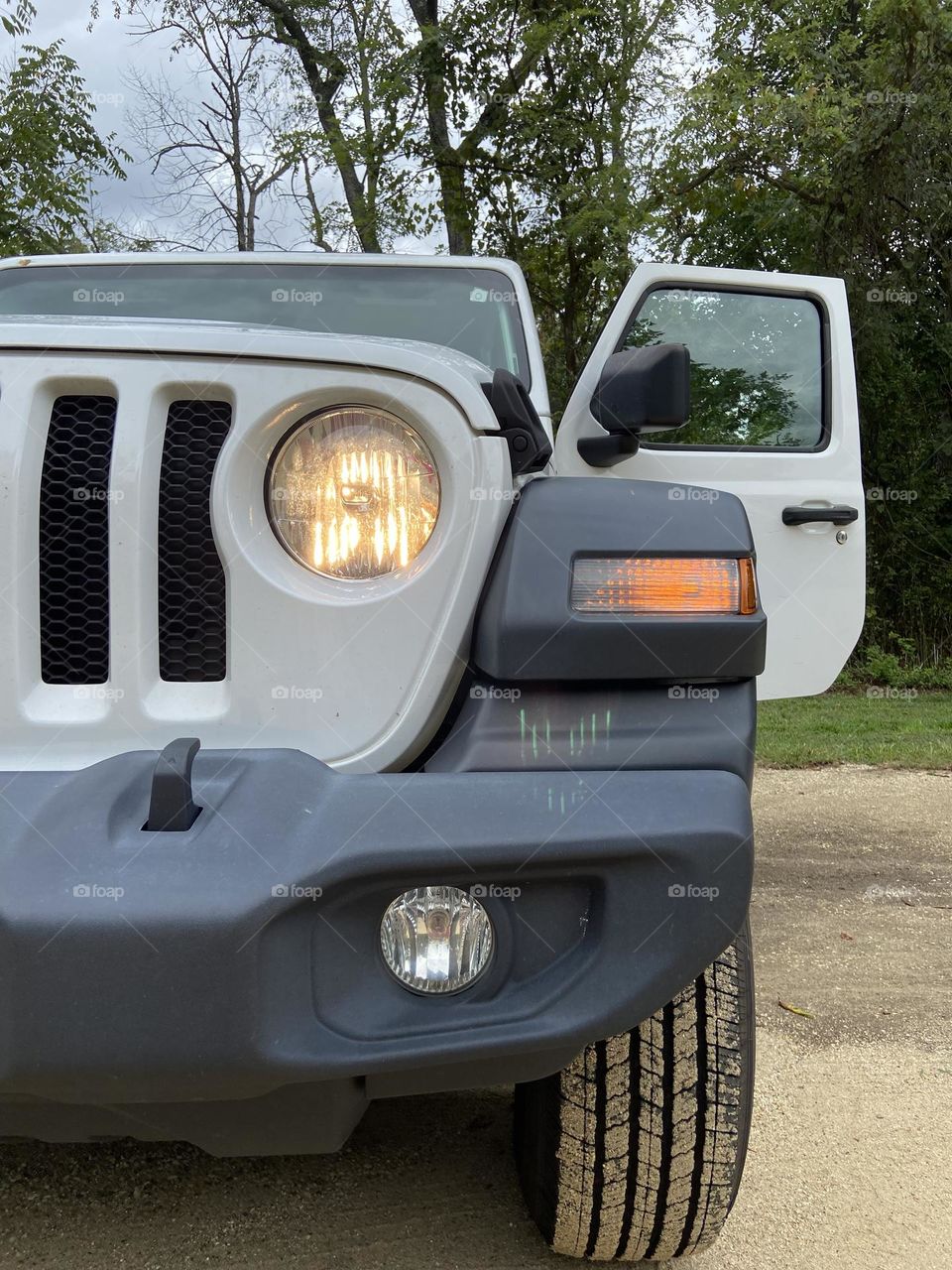 A view of the driver’s side of my 2018 Jeep Wrangler Sport. The tire is dirty from driving through the sandy ground and trees are in the background.
