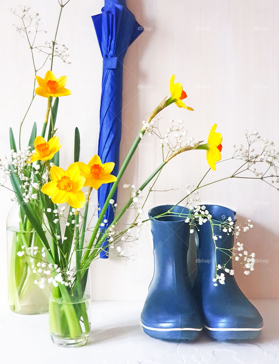 A bouquet of yellow and white spring flowers in a glass vase next to a blue umbrella and blue rubber boots on a white wooden background. Daffodils in the interior