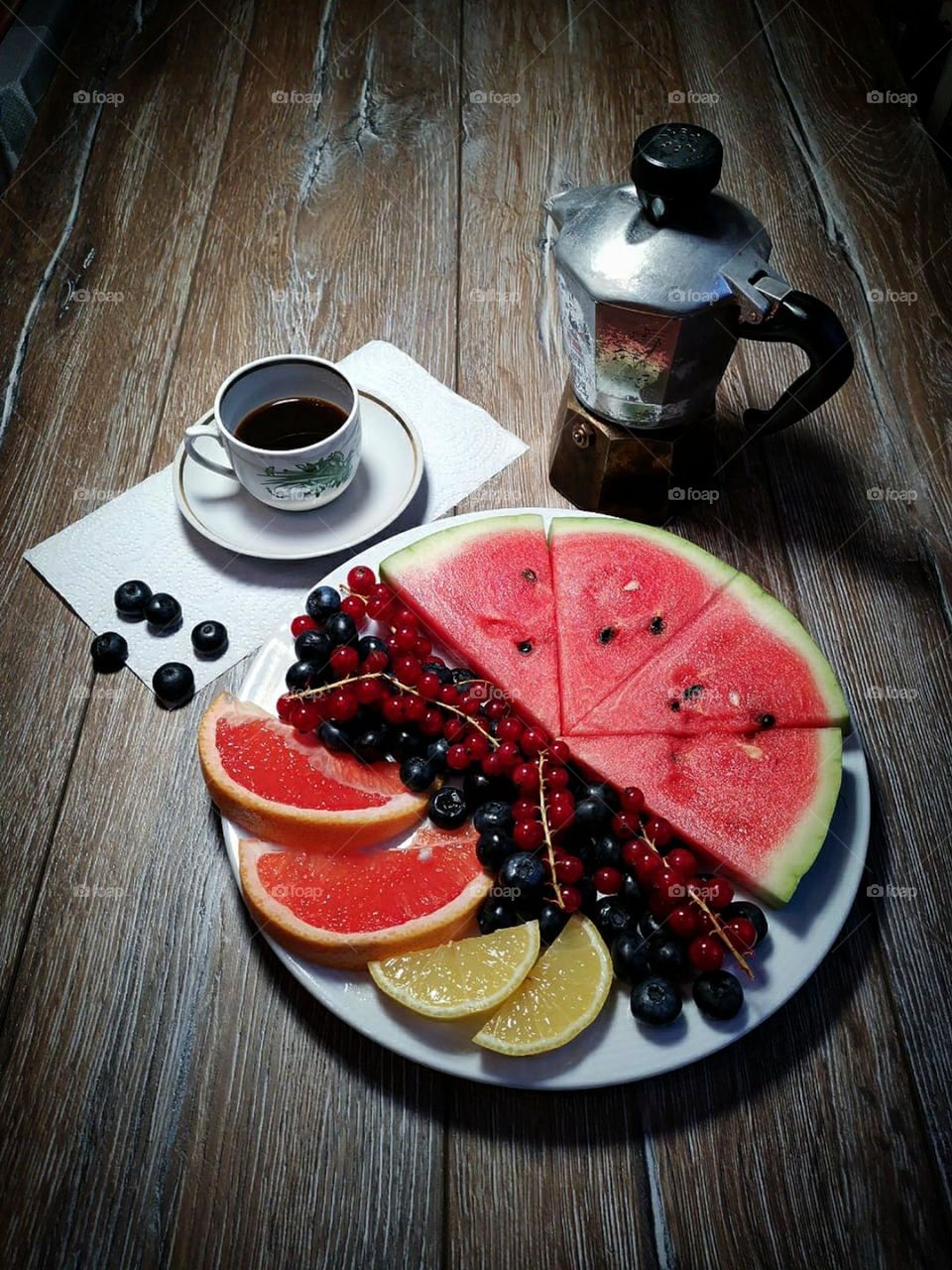 On a wooden background there is a plate with sliced pieces of red watermelon, with bunches of red currants, with black blueberries, with two slices of yellow lemon and with two slices of red grapefruit. Nearby is a cup of black coffee and a coffee