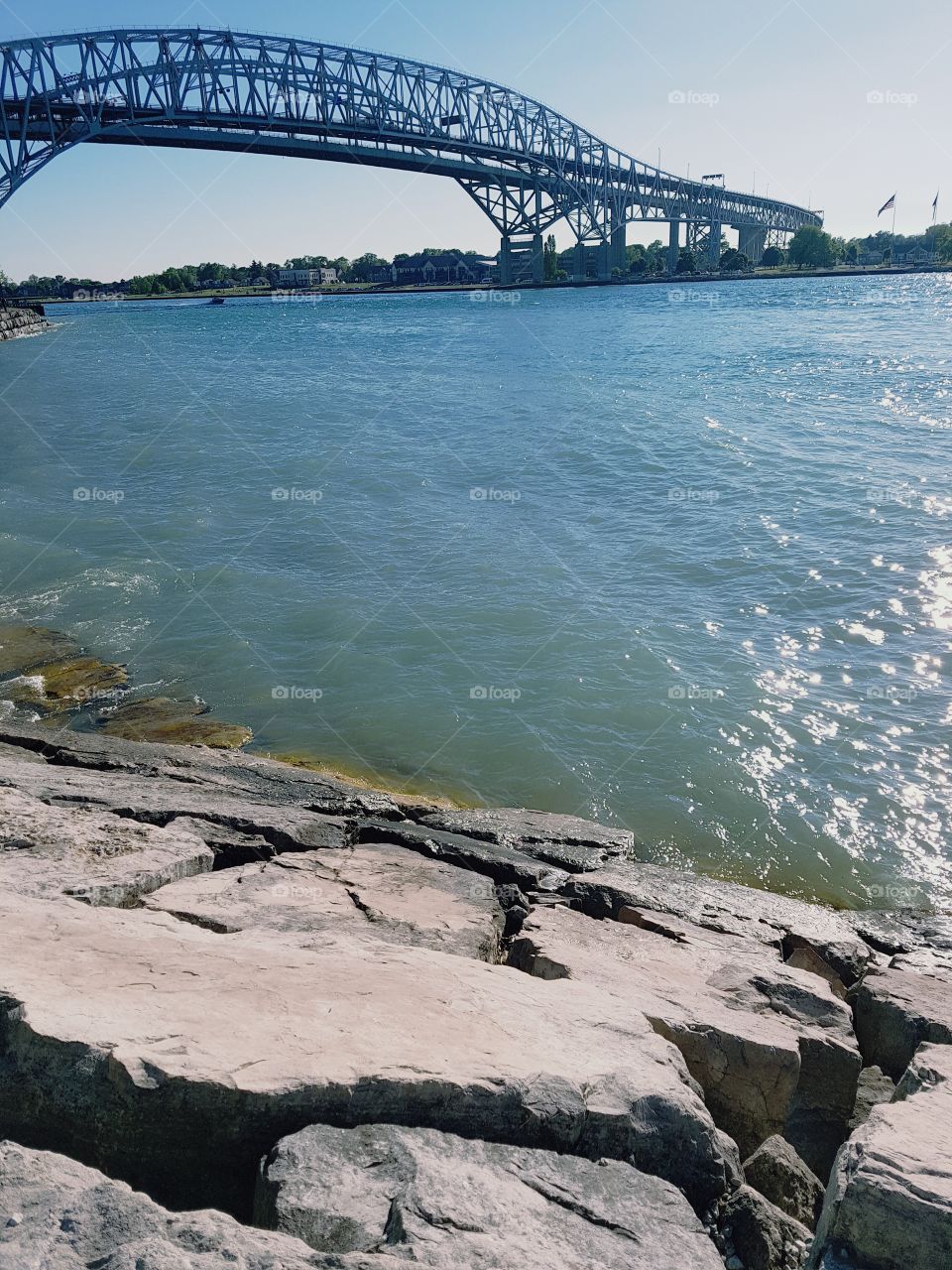 The bluewater bridge between America and Canada cascades in the background of the blue rippling water of the border, and the rocky shore.