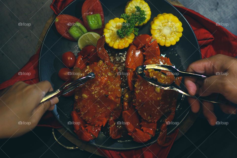 Crab flavored in creamy garlic sauce served with corn, tomatoes, lemon wedges and cherries on a black plate set on a red cloth set in a wooden setting. Asian flavor crab.