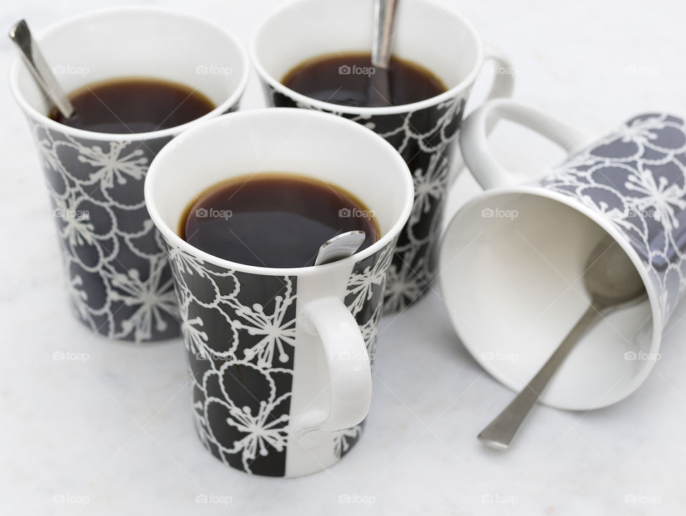 Three black and white mugs filled with strong black hot tea with spoons and one empty mug lying down.