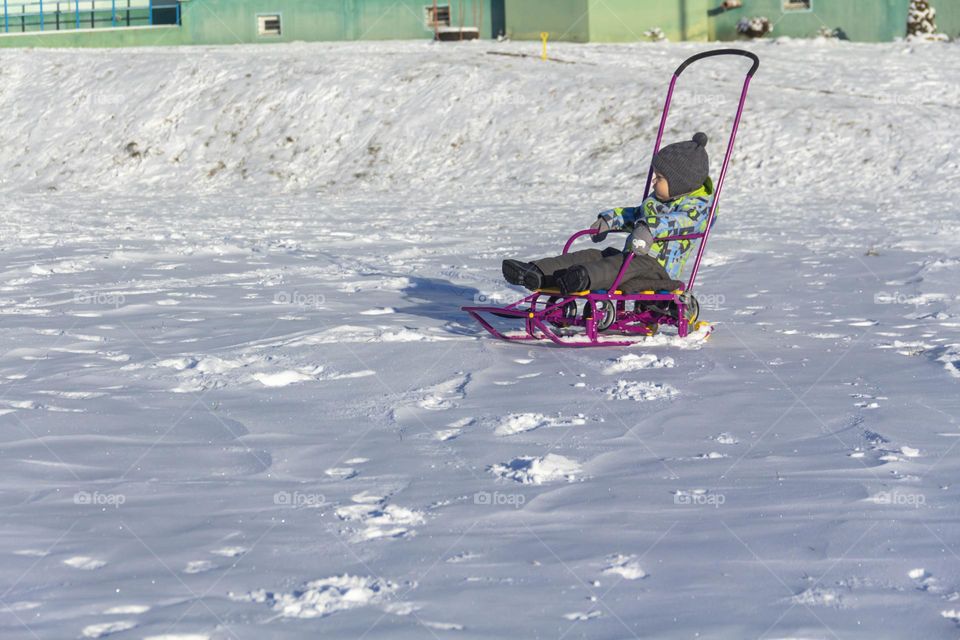 A child with a serious expression on his face in winter clothes jackets, pants, hat and boots in winter on white snow on the street and in the park in nature sledding and playing winter fun.