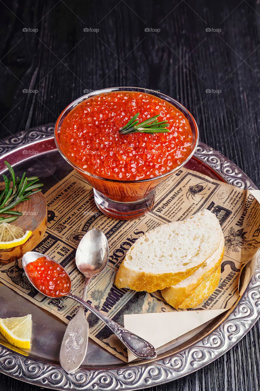 Close up of a bowl of red caviar garnished with rosemary leaves alongside slices of lemon and bread in a metal plate .