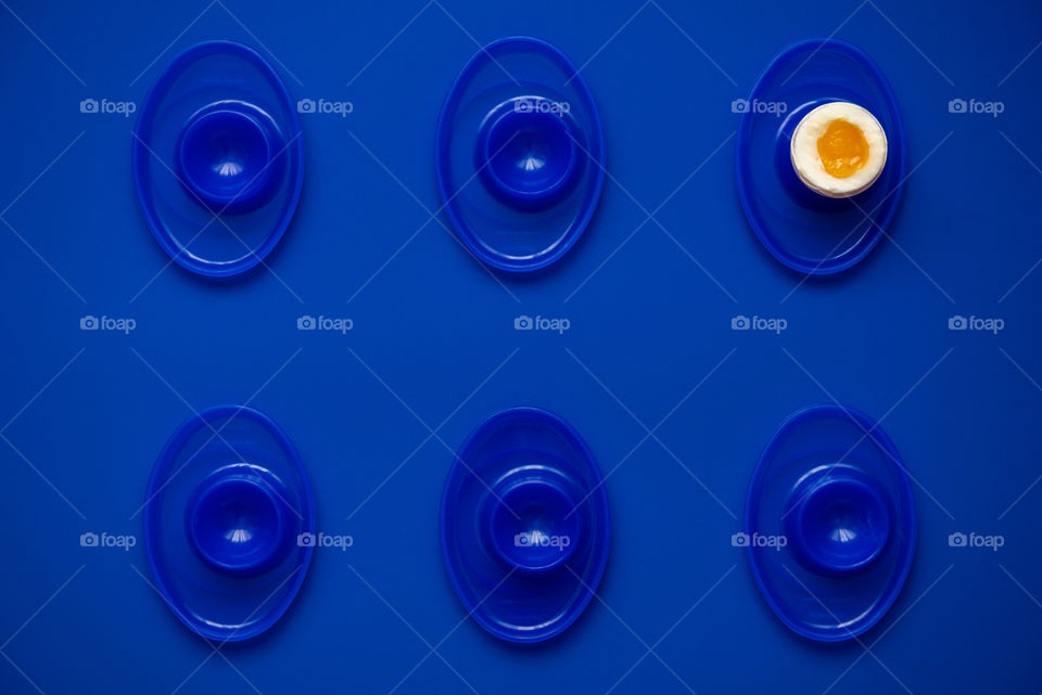 Monochromatic blue flat lay of a single soft boiled egg in an egg cup in a row of additional empty egg cups