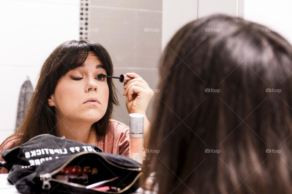 Mirror photo of young woman doing her make-up and applying mascara
