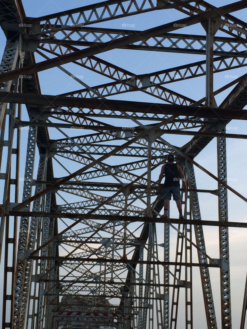 A teenage guy climbing on a steel bridge. The zig-zag steel beams interlacing in front of the light blue sky. 