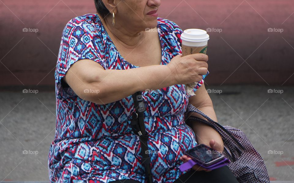 A women seated at bus stop bench with a Starbucks coffee cup in one hand and a smartphone in the other hand,