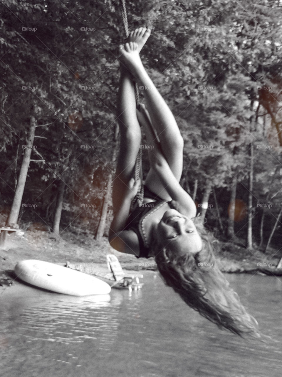 Child swinging upside down on rope swing with long hair dangling above water.