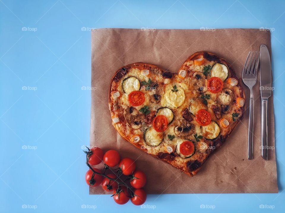 Delicious vegetarian pizza in the shape of a heart for Valentine's Day with tomatoes, vegetables and cheese on a blue background.