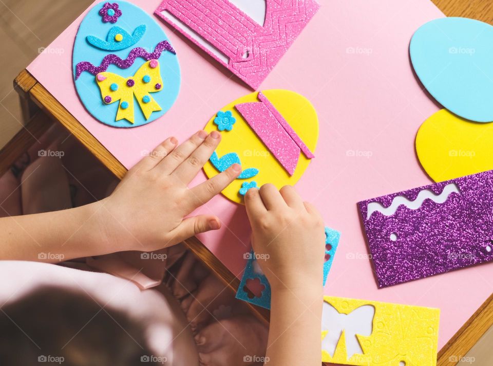 One little caucasian girl glues a blue felt flower sticker on a yellow easter egg while sitting at a children's table in the room, close-up top view. Creative kids concept, easter preparation.