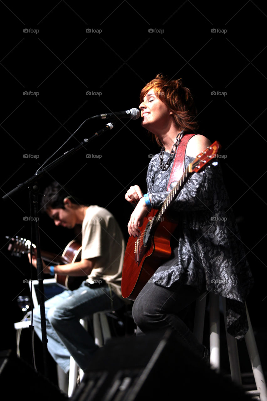 Woman sings and plays guitar with a musician man in the background 