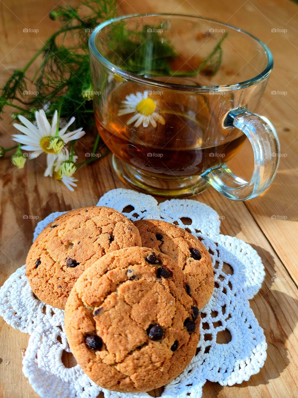 Composition. On a wooden background. White knitted napkin with oatmeal cookies with chocolate pieces; a colorless mug with chamomile tea in which a chamomile flower floats. There is a bouquet of daisies next to the mug