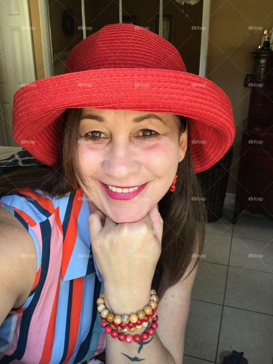 A smiling lady posing, she is wearing colorful red hat and a striped dress,red lipsticks and natural jewelry.