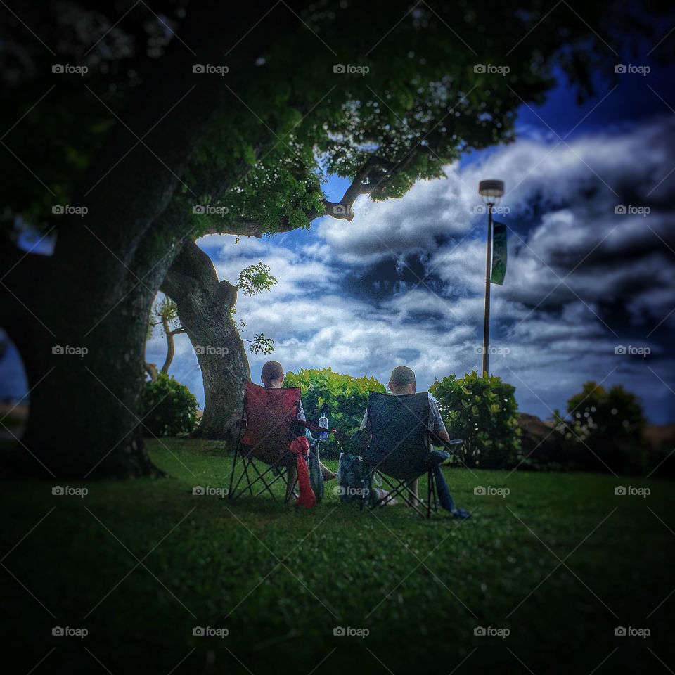 Old couple sitting in folding chairs under a tree as storm clouds gather
