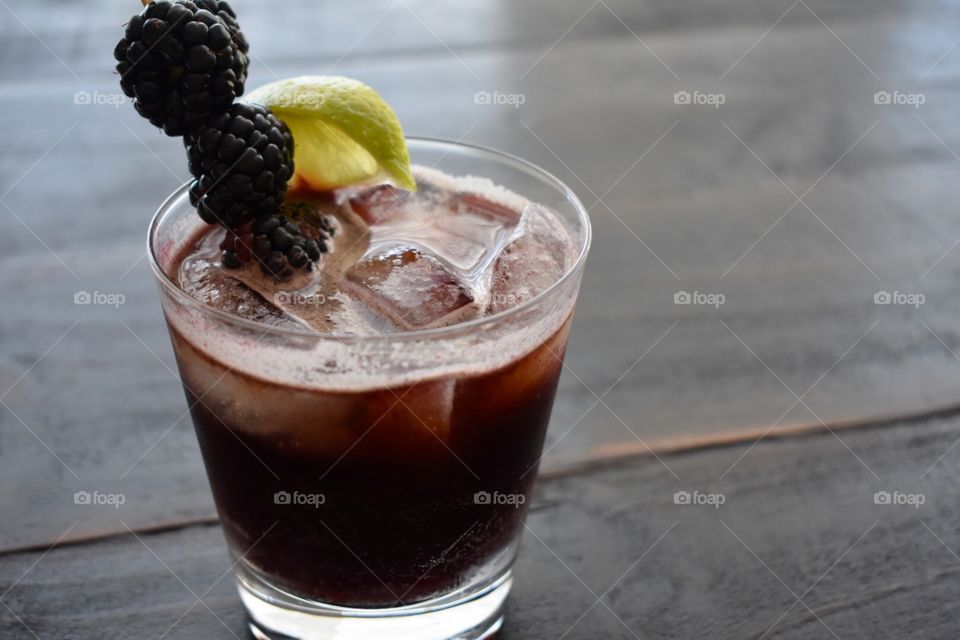 A dark purple cocktail served on the rocks in a double old fashioned glass and garnished with blackberries and a lime wedge