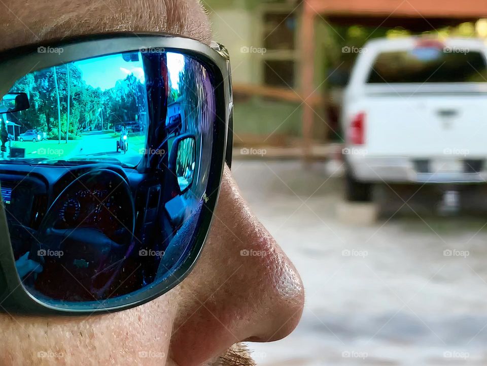 Commuting: Driver man driving with sunglasses passing vehicles seen from the left window and reflection of the road, the motorcycle and the steering wheel with his hand holding it, the interior of the pickup truck and the side mirror from his glasses