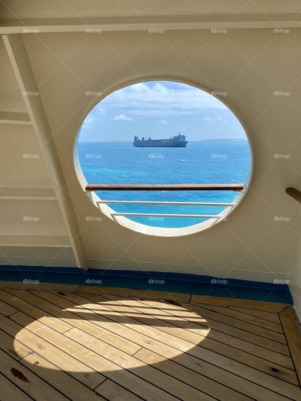 The sun shining through a porthole on the side of a cruise ship with a view of a cargo ship and the ocean