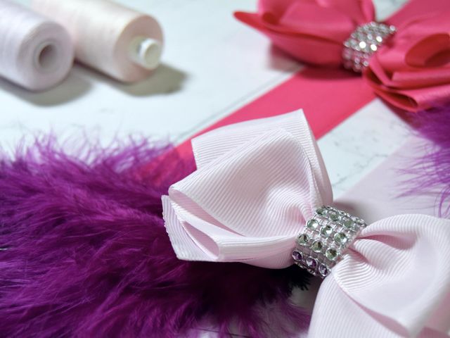 Two bows, one pale pink, the other hot pink, sit on bright pink & pale pink ribbon & a decorative bright fuchsia feather, accompanied by medium sized pale pink cotton reels on a white marble flatlay background.
