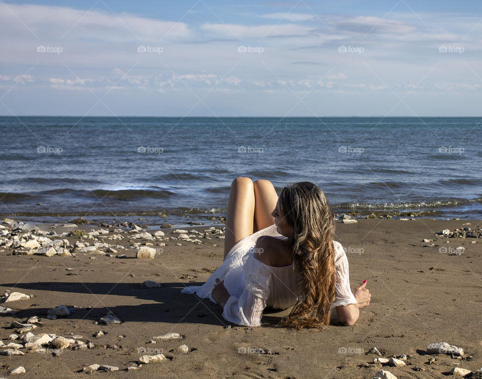 A girl with long hair is resting on the seashore at sunset of the day, back view.