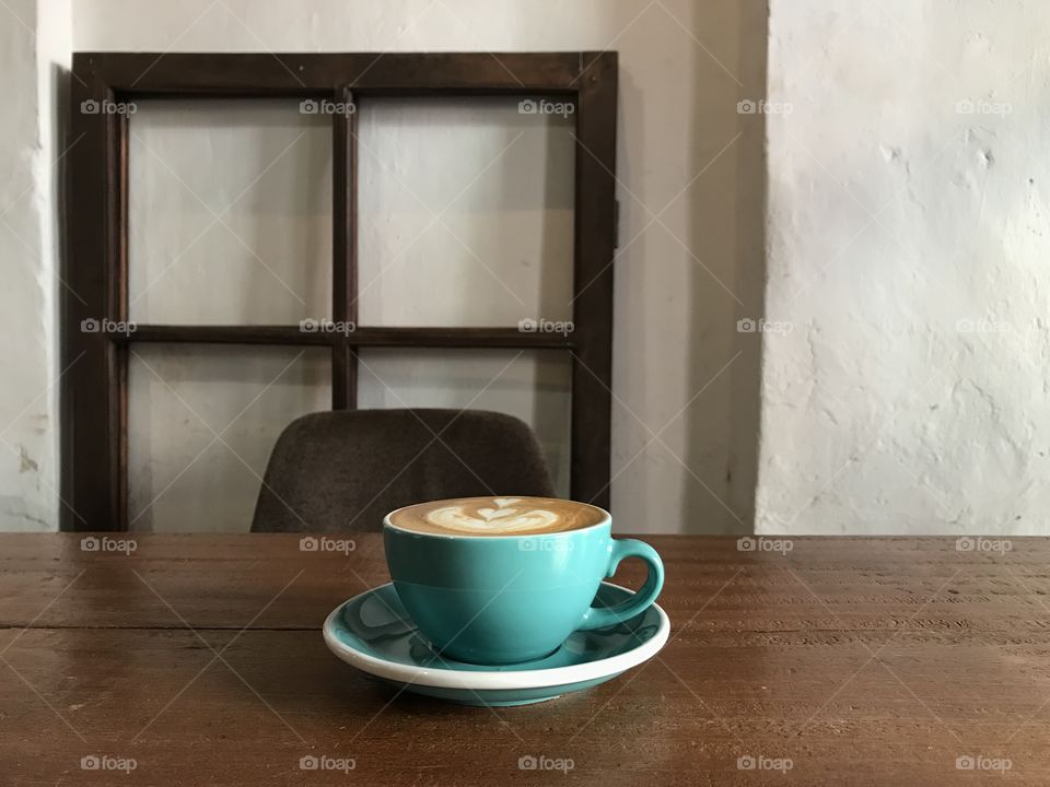pastel green cup of hot cappuccino coffee with saucer on wooden table and brown chair and antique wooden window frame as decoration with white cement wall in coffee shop