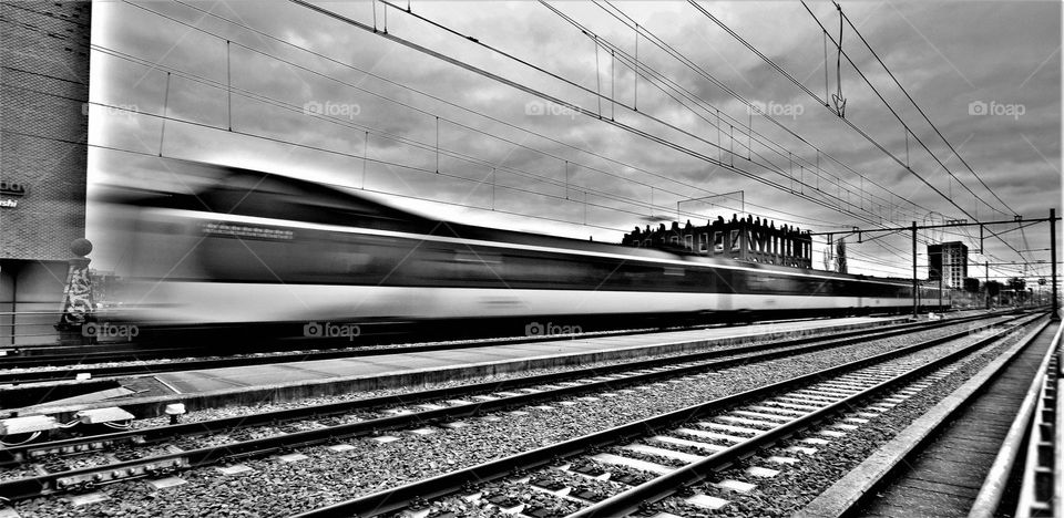 black and white image with crossing train on high speed on a railway with high voltage cables and clouds in the sky