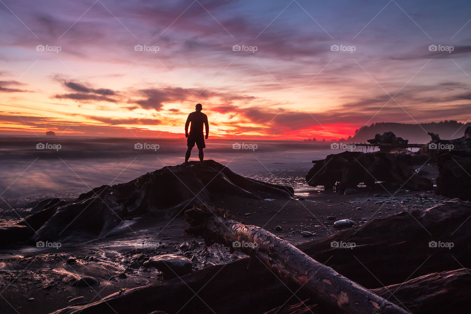 Silhouette of a young man standing on a piece of driftwood on a beach, looking off into the distance at a beautifully vibrant sunset over the ocean. 