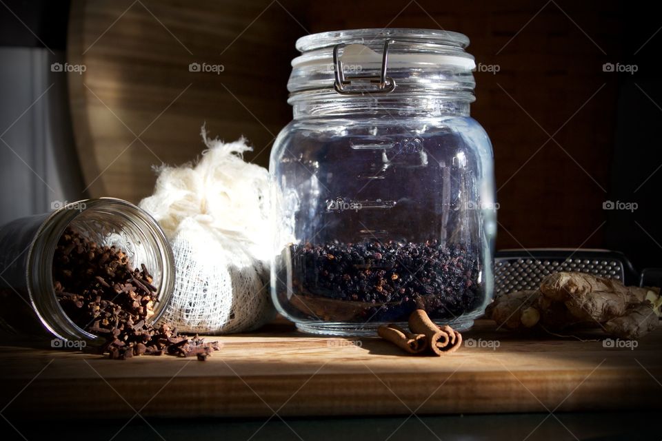 Ingredients for elderberry syrup—cloves in small glass jar, elderberries and chopped ginger in cheesecloth, dried elderberries in sealed glass jar, cinnamon sticks and ginger root with grater on wooden surface