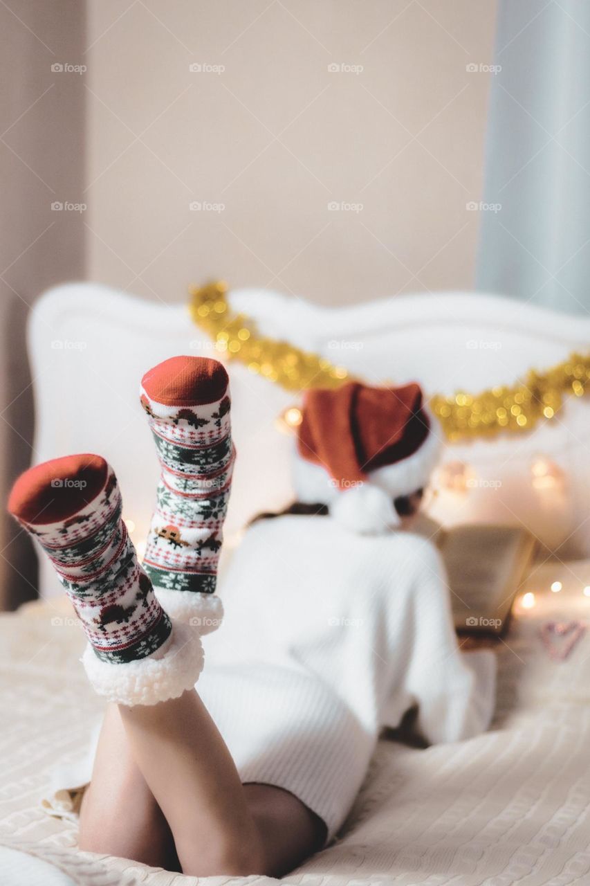 Young caucasian teenager girl in red socks christmas ornament, white sashkanlm dress and santa claus hat lies on her stomach on a wool with a garland and tinsel reading a book in anticipation of the holiday, vtd side close-up with selective focus.