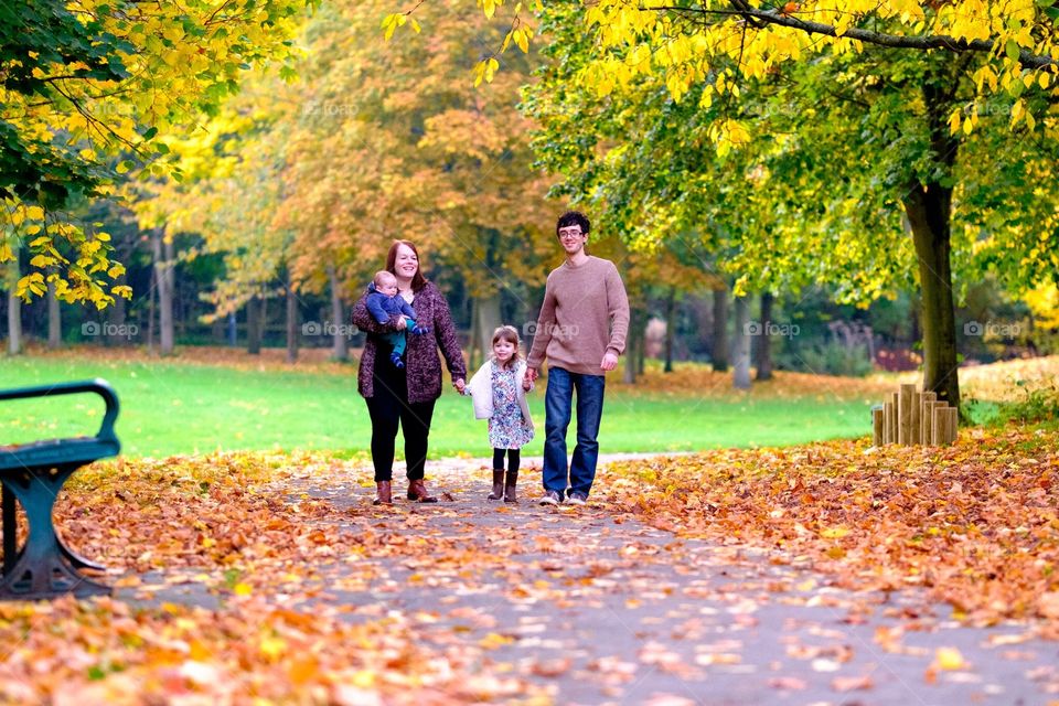 A family walk in the park. . A young family, comprising father, mother, girl aged three and baby boy, walk through a park in Autumn with fall colours