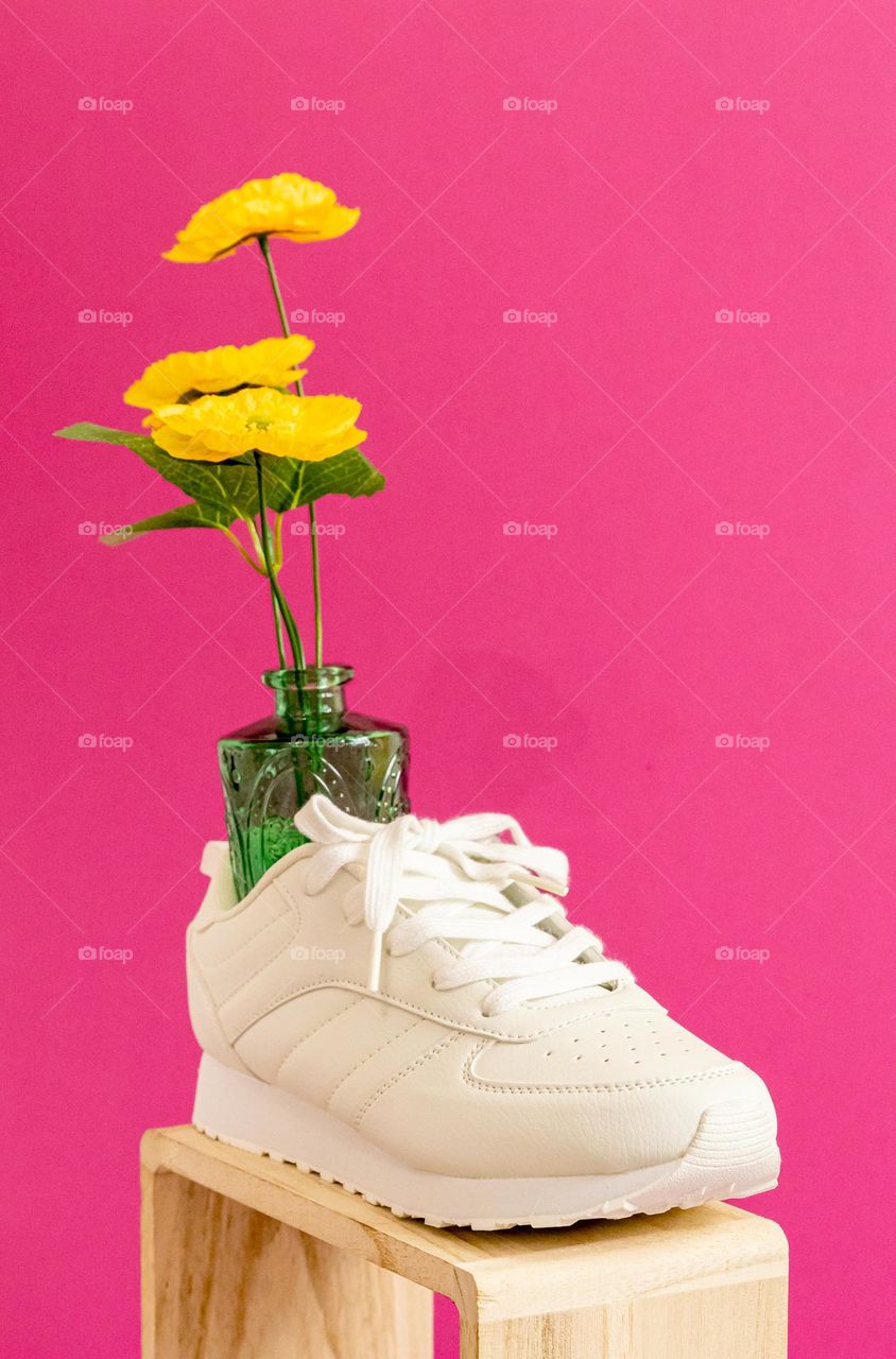 One white sneaker with a green glass vase and yellow flowers stands on a wooden box and on a bright pink background, close-up side view.