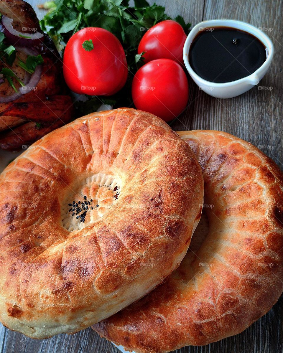 Two bread cakes (chureki) lie on a wooden background. In the background, fried chicken sprinkled with chopped onions and cilantro, red tomatoes, a bunch of cilantro and a white gravy boat with Narsharap (boiled pomegranate juice)