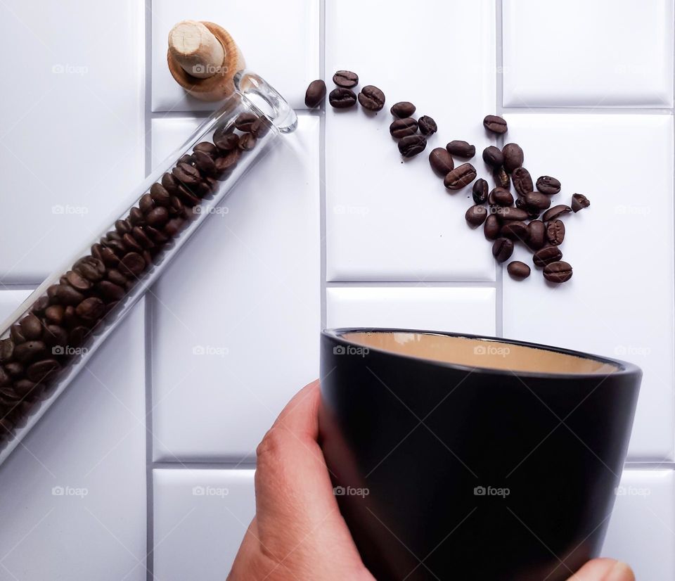 Coffee beans spilling out of slender glass container into a black coffee mug being held by a hand.  This is on a vertical white kitchen tile background.