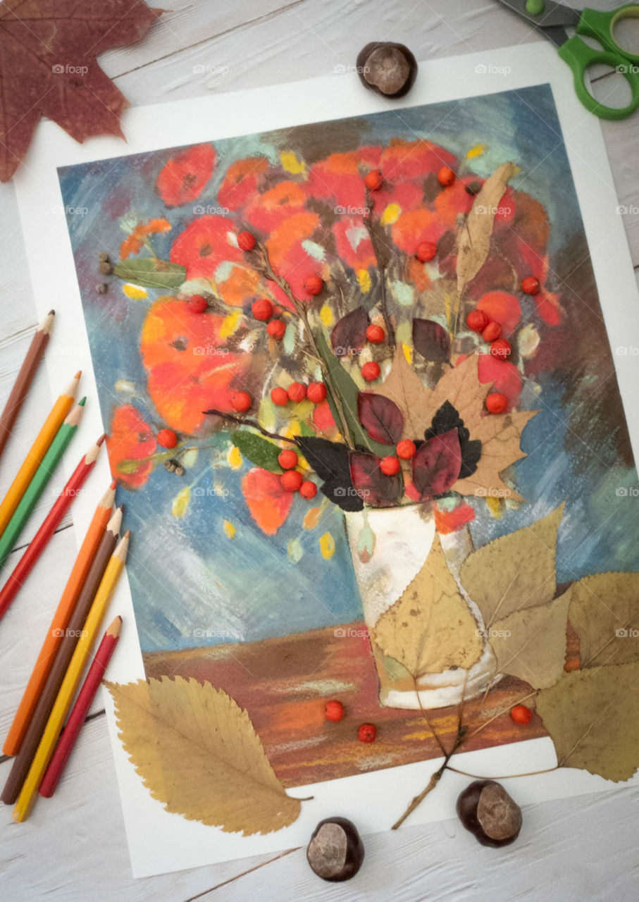 Children's creativity.  Crafts, children's painting, applique with autumn leaves and mountain ash on a light wooden background with chestnuts, multi-colored dry leaves and pencils.  Vase of flowers, dry autumn foxes, twigs and mountain ash